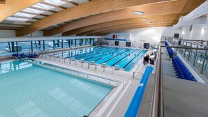Una piscina interior con varias pistas, vigas de madera en el techo y una barrera de cristal alrededor del área de la piscina.