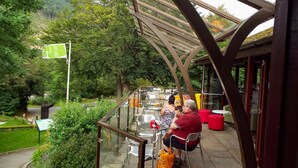 Un área de comedor al aire libre con mesas y sillas, rodeada de follaje y montañas al fondo.