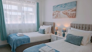 Ein Hotelzimmer mit zwei Betten, jeweils mit blauer Bettdecke und weißen Kissen. An der Wand hängt ein Gemälde mit einer Strandansicht, und es gibt ein Fenster mit Vorhängen.