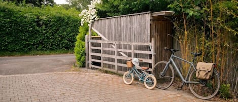 A paved driveway leading to a wooden gate, a bicycle with a bag, and a shed in a garden setting.