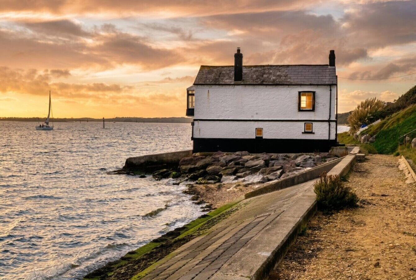 A coastal house with a stone pathway leading to the entrance, situated by the sea with a sailboat in the background.
