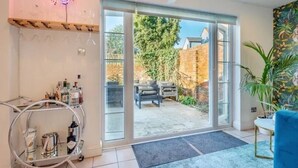 Un salon moderne avec une porte vitrée donnant sur une terrasse, un néon, un chariot en métal rempli de bouteilles et un canapé bleu.