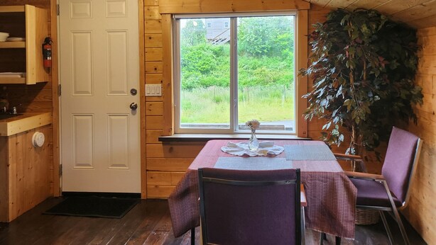 A cozy cabin interior with a wooden ceiling, a dining table with chairs, a window with a view of greenery, and a door with a fire extinguisher. - Cozy, Quite, Private Loft, Near Ocean, 
Center of Haida Gwaii (Port Clements) (Port Clements)
