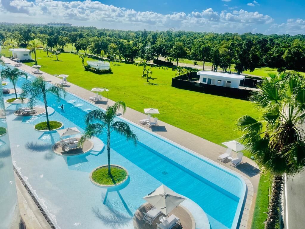 Una piscina amplia con numerosas tumbonas y palmeras, rodeada de un césped cuidado y un edificio al fondo.