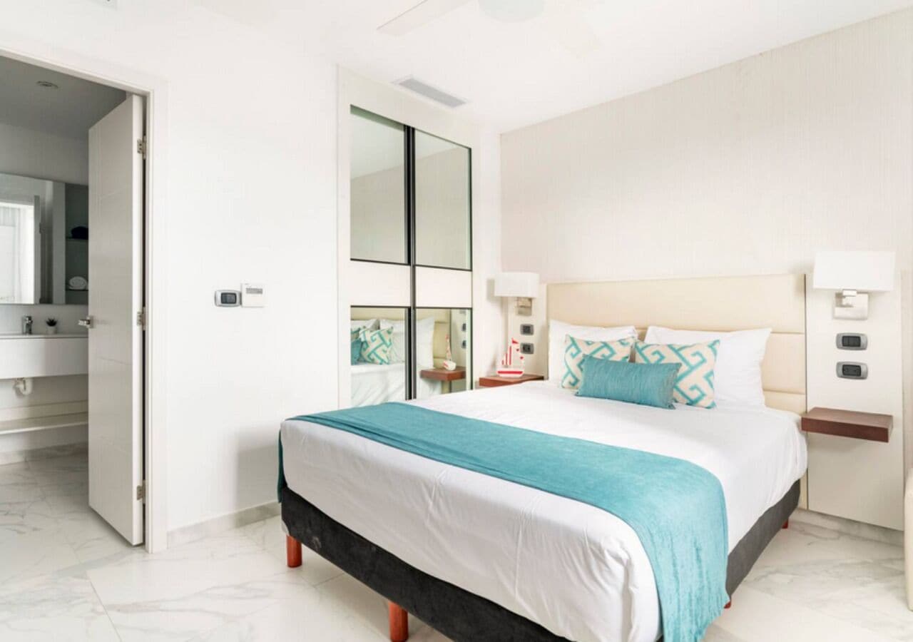Una habitación de hotel moderna con una cama grande, una mesita de noche, un espejo y un baño visible a través de una puerta abierta.