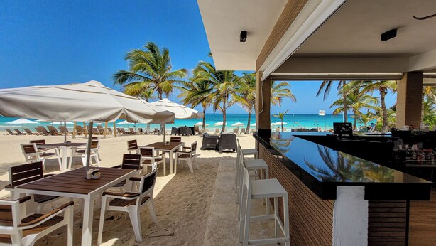 A beachside bar with white chairs and umbrellas, a pool, and palm trees. - Cana Rock Star Condo Hotel (Punta Cana)