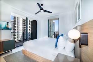 Habitación de hotel moderna con una cama grande, ventilador de techo, televisor y vista a la ciudad a través de amplios ventanales.