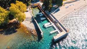 Vue aérienne d’un quai avec des bateaux, une plage et un bâtiment en pierre entouré d’arbres.