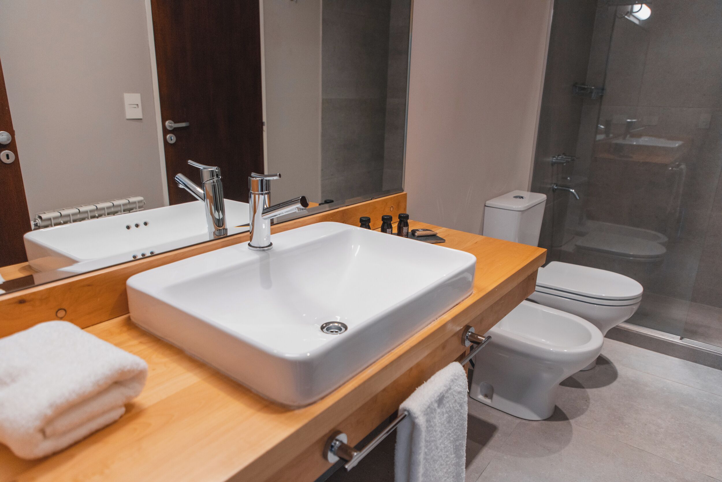 Un baño moderno con un lavabo grande blanco, un tocador de madera, un espejo, un inodoro y una cabina de ducha de vidrio.