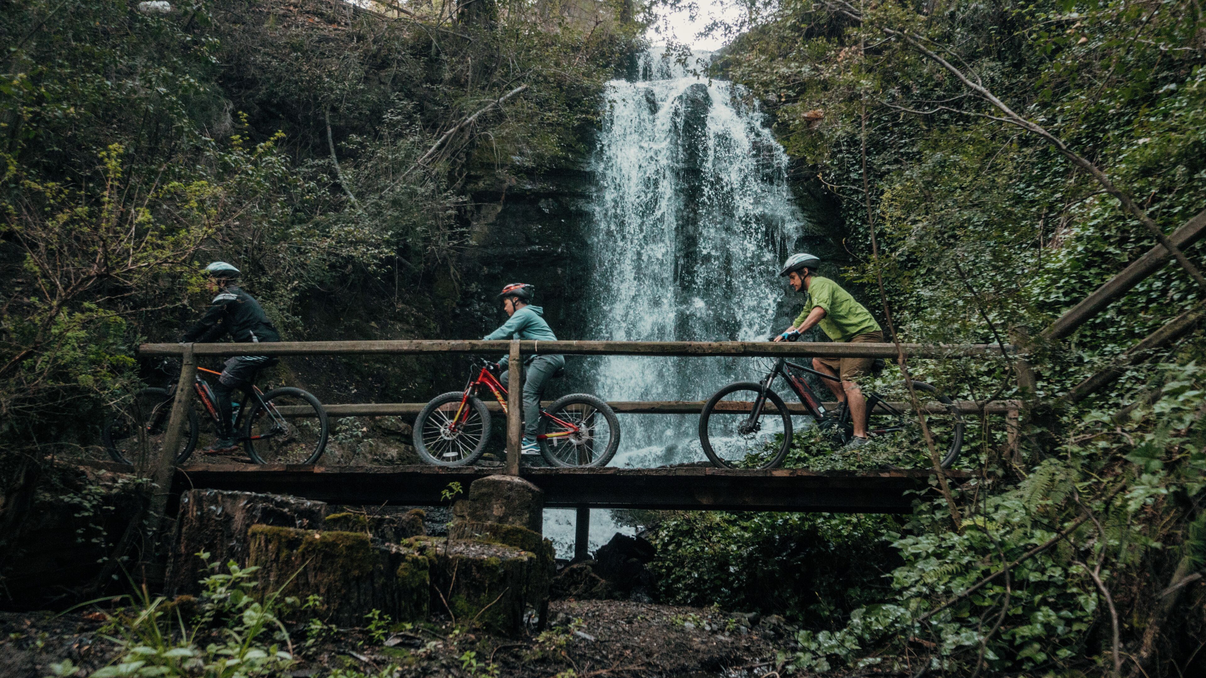 Trois cyclistes sur un pont en bois surplombant une cascade dans une zone boisée.
