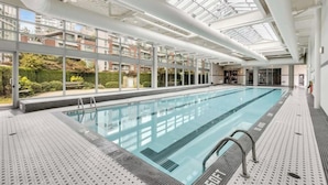 A modern indoor swimming pool with clear glass ceilings, a tiled floor, and a view of residential buildings outside. - Beautiful Garden View Pool+Hot Tub+Sauna/Large Free Parking (Vancouver)