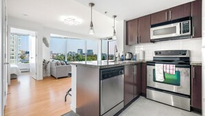 A modern kitchen with stainless steel appliances, a dishwasher, and a large window overlooking a cityscape. - Beautiful Garden View Pool+Hot Tub+Sauna/Large Free Parking (Vancouver)