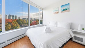 A modern hotel room with a large window offering a city view, a neatly made bed, and a small bedside table with a plant. - Beautiful Garden View Pool+Hot Tub+Sauna/Large Free Parking (Vancouver)