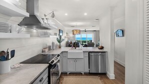 A modern kitchen with white cabinetry, a stainless steel range hood, a built-in oven, and a dishwasher. There is a large window providing a view of the ocean.