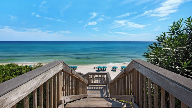 A wooden staircase leading to a beach with turquoise water, a clear sky, and beach umbrellas.