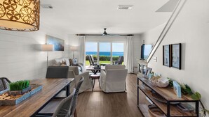 A modern living room with a dining area, large windows with a view of the ocean, and a ceiling fan.