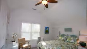 A bedroom with a bed, a chair, a ceiling fan, a window, and a wall-mounted lamp.