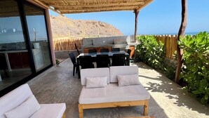 Uma área externa com mesa de madeira e dois sofás, churrasqueira e vista para o mar.