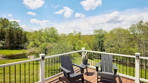 Eine Terrasse mit zwei Adirondack-Stühlen, einem Blumentopf und Blick auf einen Teich und Bäume.
