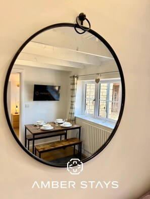 A dining area with a table set for four, a television above the table, and a window with curtains. - Four Counties Cottages, Collyweston (Collyweston)