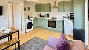 A compact kitchen with a washing machine, oven, and a dining area with a table and bench. - Four Counties Cottages, Collyweston (Collyweston)