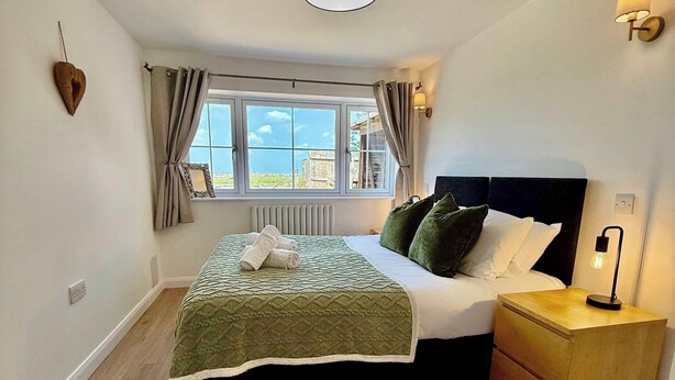 A bedroom with a bed, a nightstand, a wall-mounted lamp, a ceiling light, a window with curtains, and a heart-shaped decoration on the wall. - Four Counties Cottages, Collyweston (Collyweston)