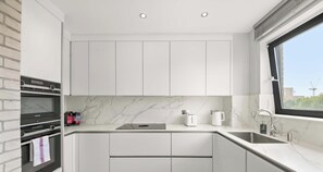 A modern kitchen with white cabinets, a marble countertop, a built-in oven, and a window with a view. - Lawrence Court, Acton, London (Greater London)