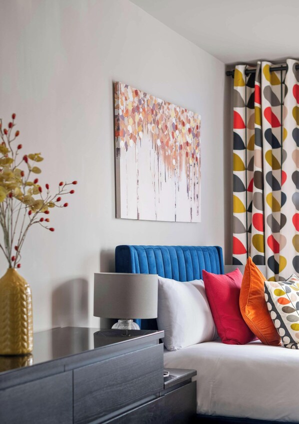 A modern bedroom with a blue headboard, a bedside table with a lamp, and colorful pillows. - Lawrence Court, Acton, London (Greater London)