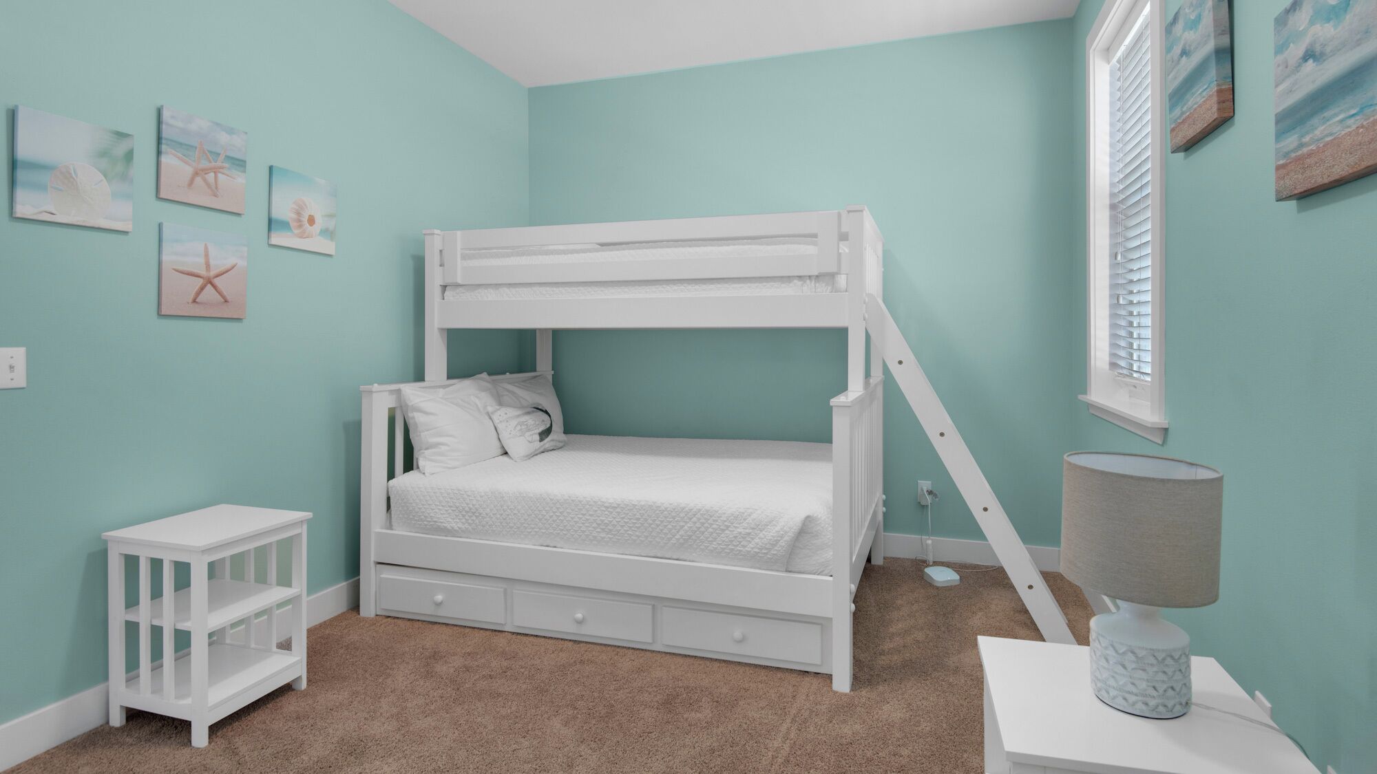 A bunk bed room with a teal wall, a small white nightstand, a lamp, and starfish-themed artwork.