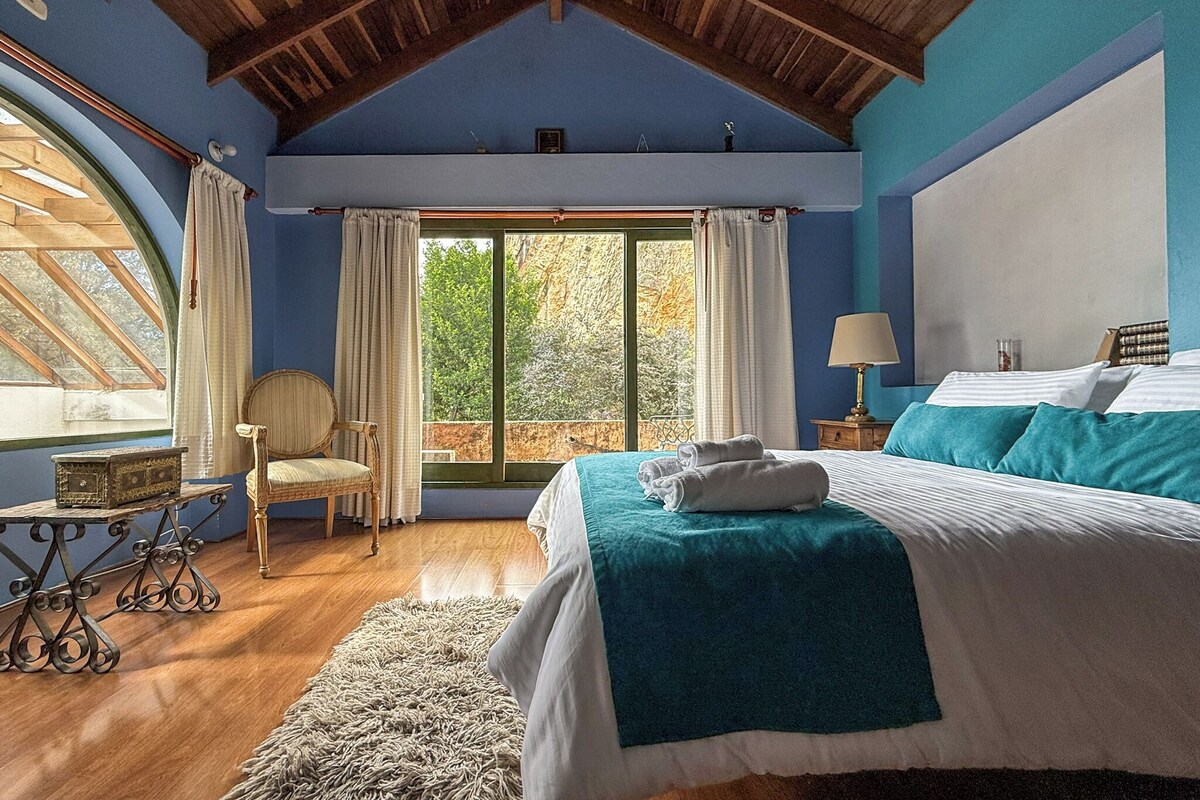 A bedroom with a large bed, a chair, a table, and a view of trees through a window.