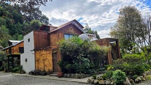 Una casa rústica amb un teulat inclinat, una finestra gran i un jardí amb diferents plantes i una banca.