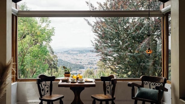 Habitació amb una finestra gran que ofereix una vista panoràmica d'arbres i un paisatge urbà a la distància. Hi ha una taula petita amb una planta i una tetera, i dues cadires amb coixins.