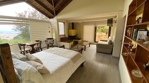 Un dormitorio con una cama grande, un sofá, una mesita y un televisor. Hay ventanas con persianas, una estantería y vistas al exterior.