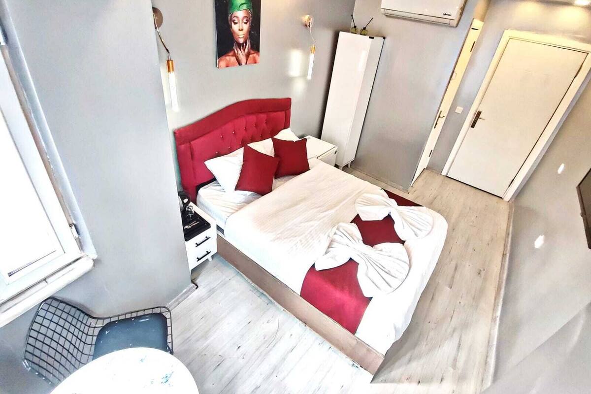 Une chambre avec une tête de lit rouge, un lit avec une literie blanche et rouge, une petite table de chevet, un climatiseur fixé au mur et une porte.
