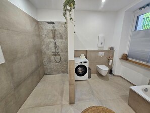 Un baño moderno con ducha, lavadora, inodoro y una ventana con persianas.