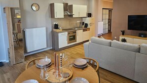 A modern kitchen with white cabinets, a microwave, and a stove. There is a dining area with a wooden table and chairs, and a grey sofa in the living room.