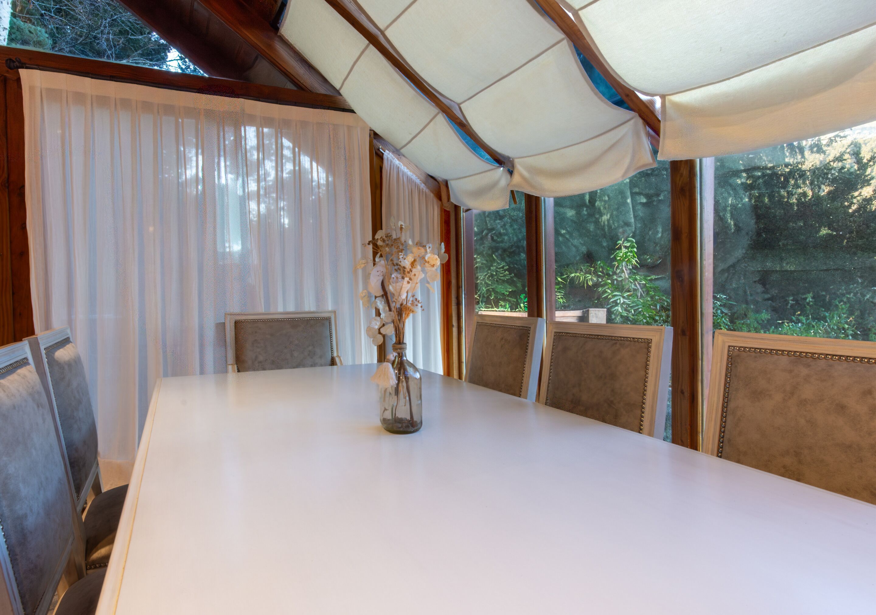 Un coin repas avec une table blanche, des chaises, un vase de fleurs et de grandes fenêtres avec des rideaux.