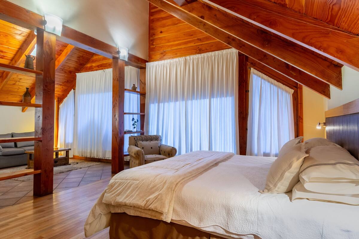 Un dormitorio con techo de madera, una cama con ropa de cama blanca, una silla y una ventana con cortinas.