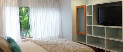 Un dormitorio con una cama, un televisor en un estante y una ventana con cortinas.