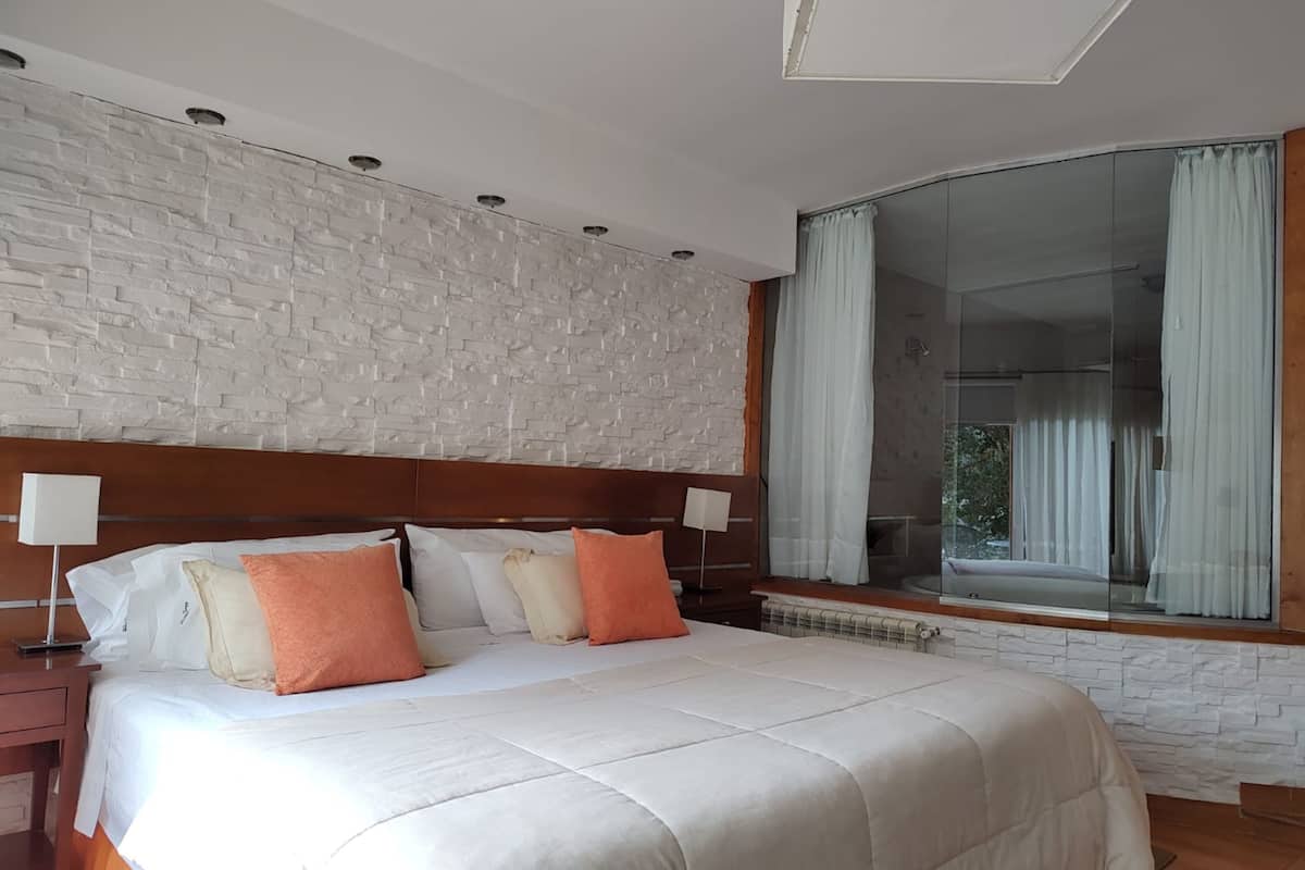 Una habitación de hotel moderna con una cama grande, un cabecero de madera, pared con textura de piedra, un ventanal con cortinas y una mesita de noche con lámpara.