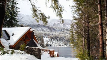 A cabin with a stone foundation and wooden exterior, situated by a lake with snow-covered mountains in the background.