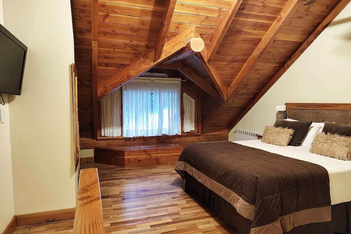 Un dormitorio con techo inclinado, suelo de madera, cama con colcha marrón y ventana con cortinas traslúcidas.