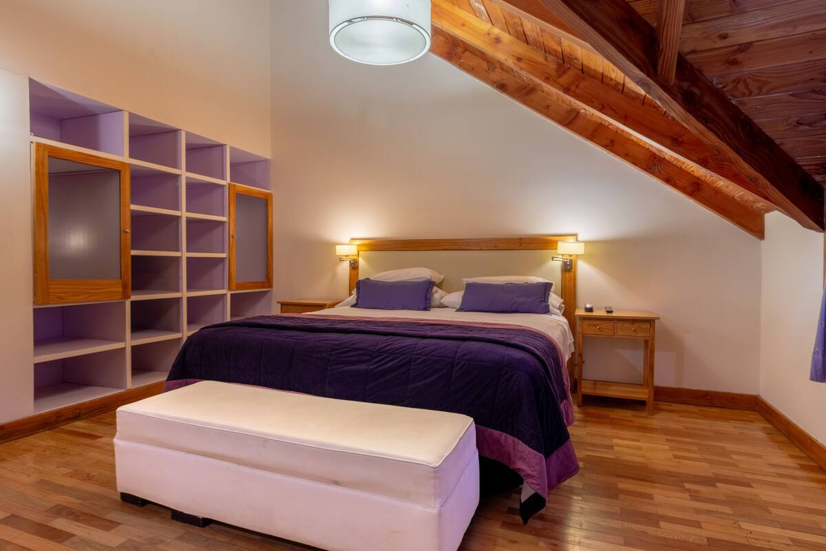 Un dormitorio con techo de madera, una cama con ropa de cama púrpura, un banco blanco y una mesita de noche de madera.