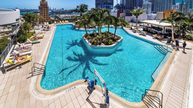 Una piscina grande con una isla de palmeras, tumbonas y un horizonte urbano al fondo.