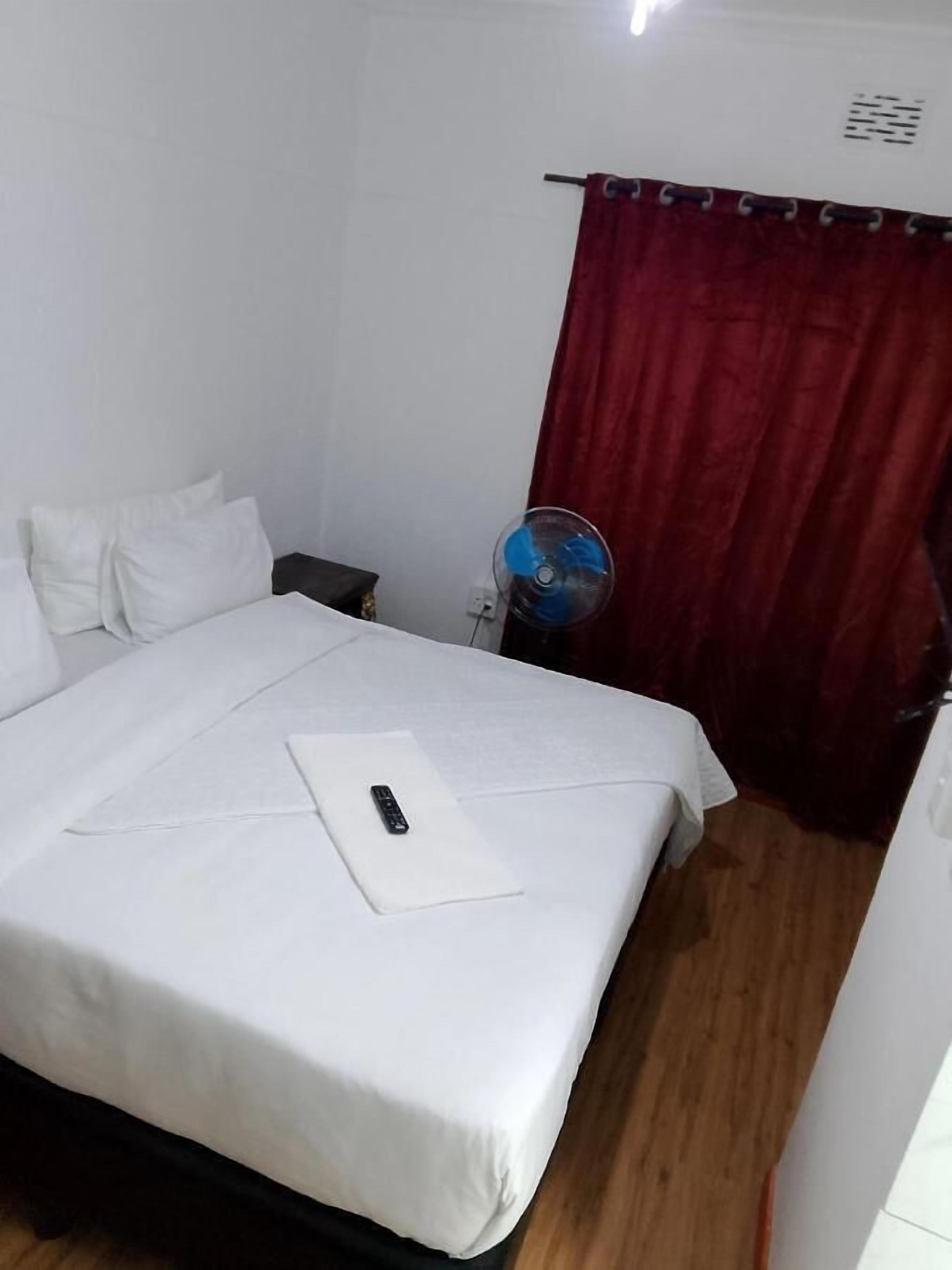 A hotel room with a single bed, white bedding, a remote control, a red curtain, and a standing fan.