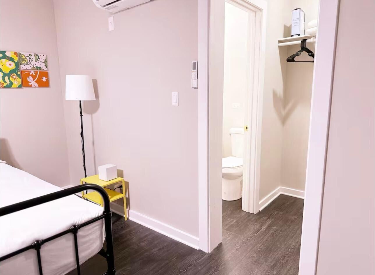A bedroom with a bed, bedside table, lamp, and a small yellow cabinet. There is a door leading to a bathroom with a toilet and a shelf with items.