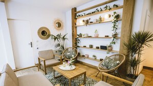 A cozy living room with a wooden coffee table, two armchairs, a sofa, a patterned rug, and a wall-mounted shelf with plants and decorative items.