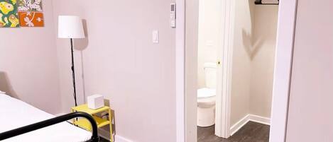 A bedroom with a bed, bedside table, lamp, and a small yellow cabinet. There is a door leading to a bathroom with a toilet and a shelf with items.