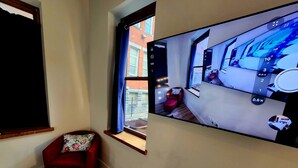 Une pièce avec un grand téléviseur fixé au mur, qui affiche les images d’une caméra de surveillance, un fauteuil rouge et une fenêtre donnant sur l’extérieur.