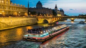 Un barco de crucero fluvial con pasajeros, un puente de piedra y edificios históricos al fondo.